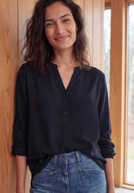 Glimmende vrouw in zwarte blouse leunt ontspannen tegen een houten wand bij een raam.