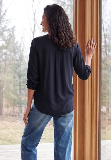 Vrouw in spijkerbroek en zwart shirt, met de rug naar de camera, kijkt uit een raam.