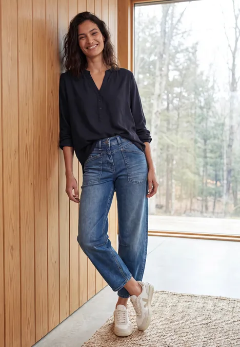 Jonge vrouw glimlachend tegen houten wand, donker blouse, spijkerbroek en witte sneakers, voor groot raam.
