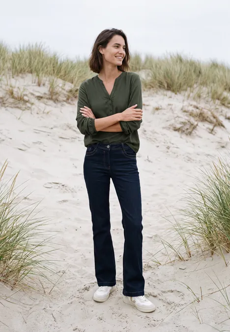 Vrouw in groen shirt en spijkerbroek, glimlachend met armen over elkaar op zandduinen.
