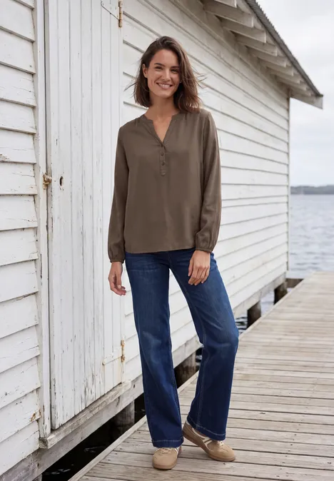 Een glimlachende vrouw in een olijfkleurige blouse en spijkerbroek staat op een houten steiger naast een wit bootshuis.