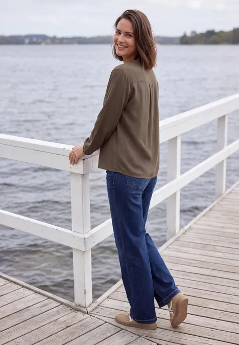 Vrouw in groene jas en blauwe jeans staat op een steiger aan het water en kijkt glimlachend over haar schouder.