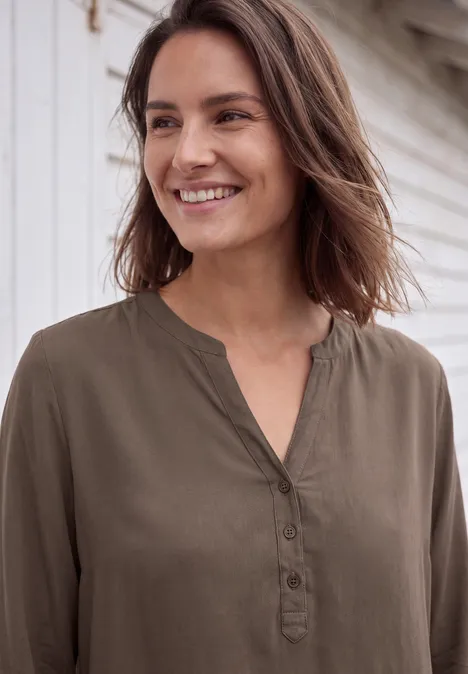 Een vrouw met halflang bruin haar draagt een olijfgroene blouse en lacht voor een lichte houten achtergrond.