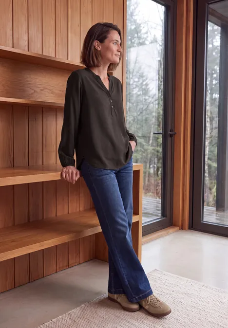 Frau lehnt an einem Holzregal und blickt aus dem Fenster eines modernen Raums.
