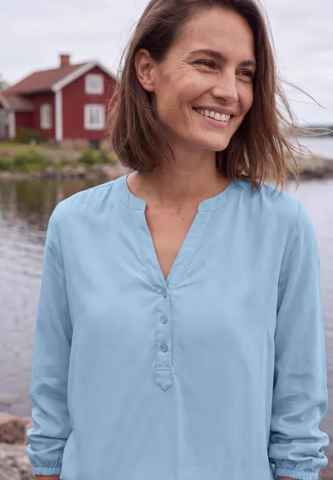 Een lachende vrouw in een blauwe blouse voor rode huisjes aan het water.