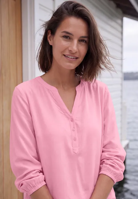 Een glimlachende vrouw in een roze blouse voor een huis, ontspannen kijkend naar de camera.