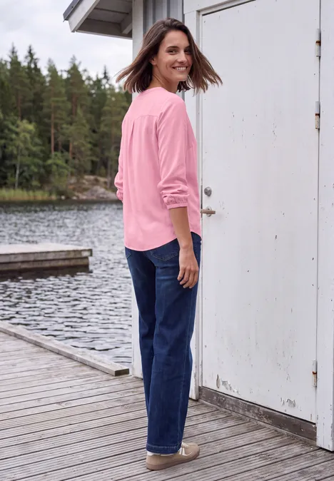 Een vrouw met een roze trui en spijkerbroek staat op een steiger bij een meer en kijkt glimlachend om.