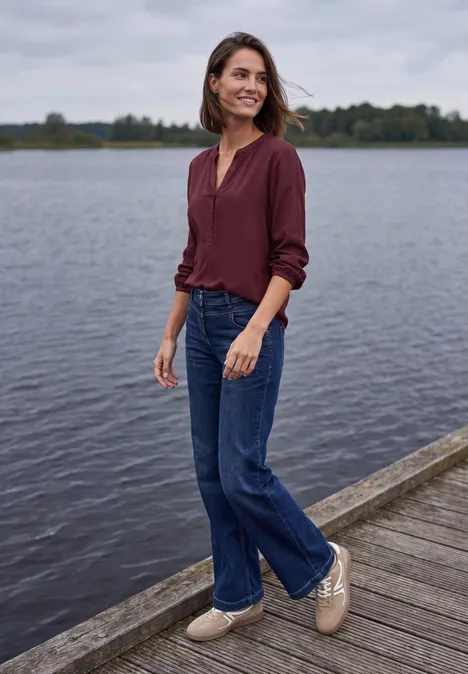 Een vrouw staat ontspannen op een steiger bij het meer, met een bordeauxrode blouse, jeans en lichte schoenen.