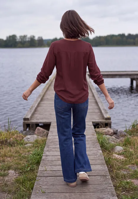 Vrouw in rood overhemd en spijkerbroek loopt over een houten steiger naar een meer, omringd door gras en bomen.
