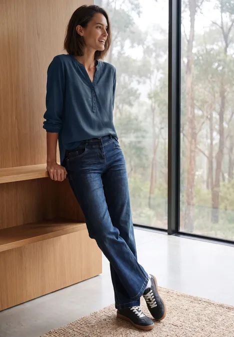 Langarm Tunika mit Split Neck moonlight blue Frau lehnt entspannt an Holzregal in modernem Raum mit bodentiefem Fenster, trägt blaue Bluse und Jeans.