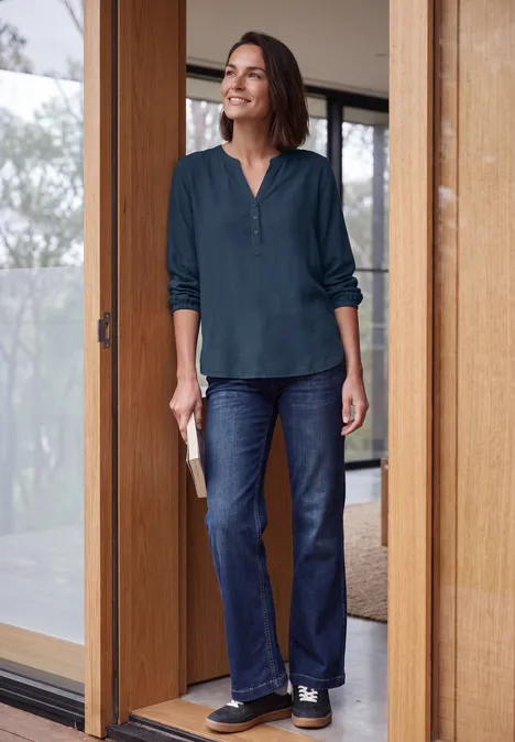 Langarm Tunika mit Split Neck moonlight blue Eine lächelnde Frau in Jeans und grüner Bluse steht im Türrahmen und hält ein Buch.