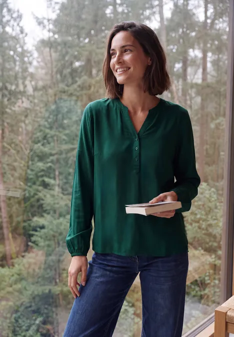 Langarm Tunika mit Split Neck dark fir green Frau in grüner Bluse steht lächelnd vor einem Waldpanorama, hält ein Buch und einen Stift in der Hand.
