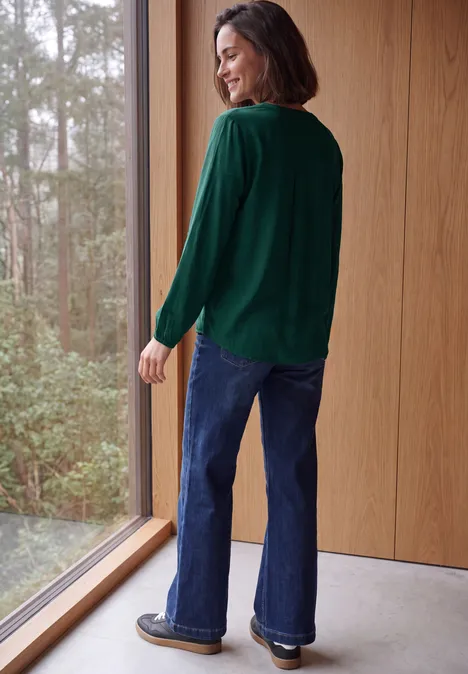 Langarm Tunika mit Split Neck dark fir green Frau steht lächelnd an großem Fenster in einem Holzraum, trägt grünen Pullover und blaue Schlagjeans.