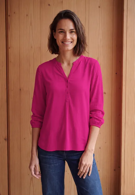 Een glimlachende vrouw in een roze blouse en spijkerbroek voor een houten wand.