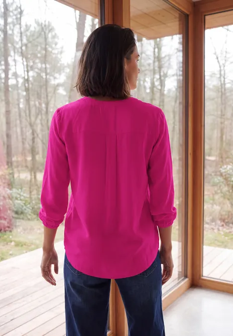 Vrouw van achteren in een felroze blouse en spijkerbroek, voor een glazen deur met boszicht.