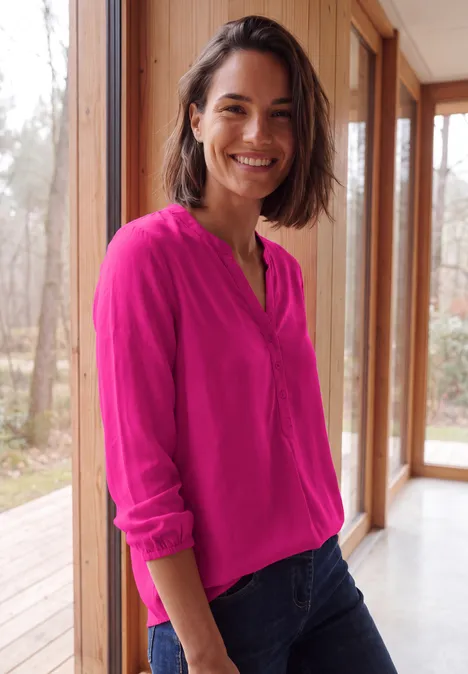 Een glimlachende vrouw in een felroze blouse en spijkerbroek staat in een lichte kamer met houten wanden.