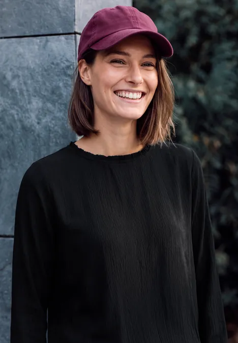 Eine junge Frau mit bordeauxroter Cap und schwarzem Oberteil lächelt im Freien.