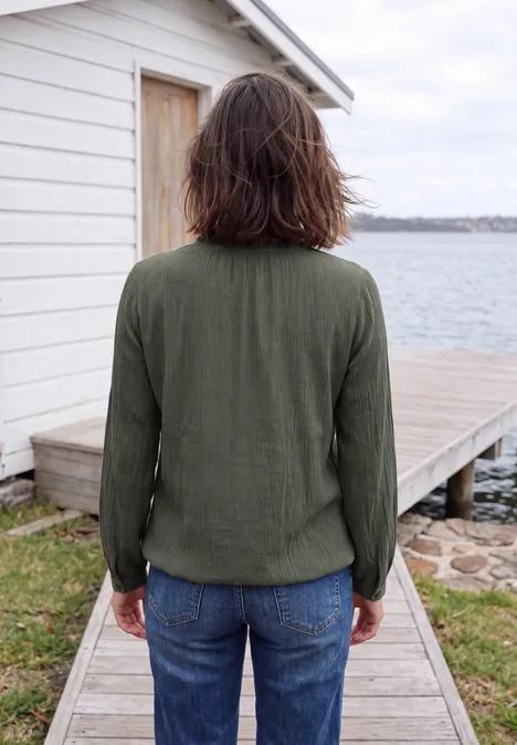 Person in grüner Jacke und Jeans steht auf einem Bohlenweg vor einer weißen Hütte am Wasser.