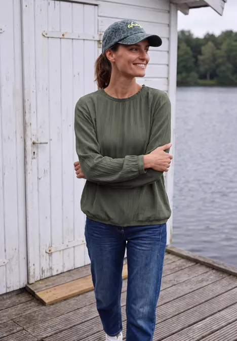 Frau mit Schirmmütze und grünem Longsleeve lehnt mit verschränkten Armen an Steg vor Seehütte.