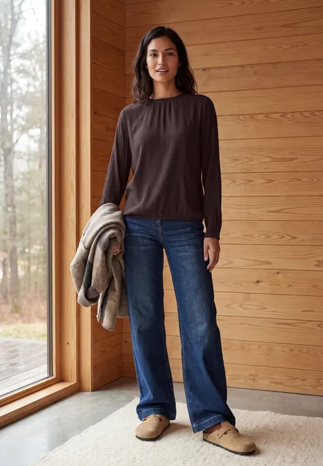 Une femme en haut marron et jean, debout dans un intérieur en bois, tenant un manteau et regardant la caméra.