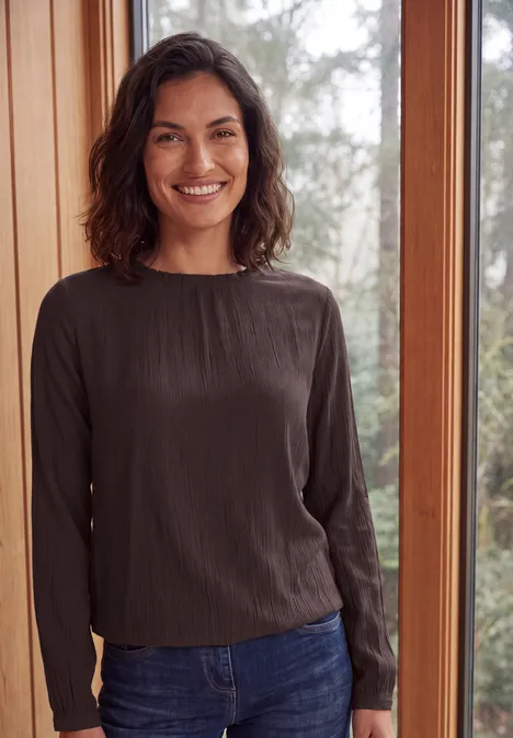 Femme souriante en haut à manches longues et jean, debout devant une grande fenêtre donnant sur la verdure.