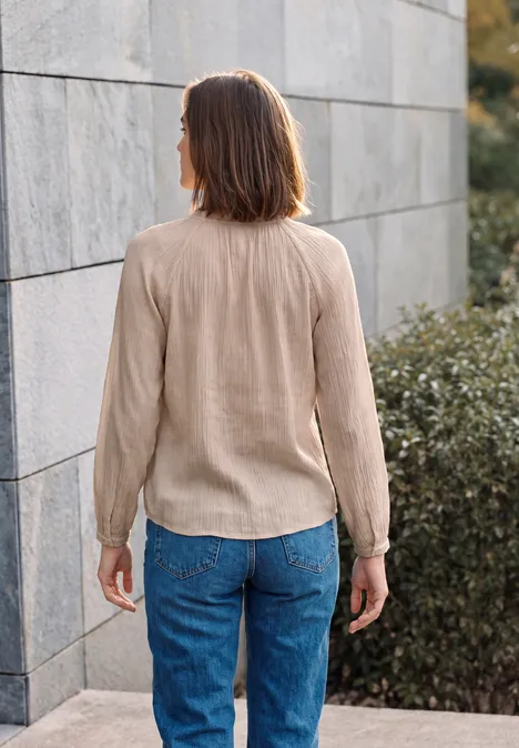 Femme de dos, vêtue d'un haut beige et d'un jean bleu, debout devant un mur et des buissons.