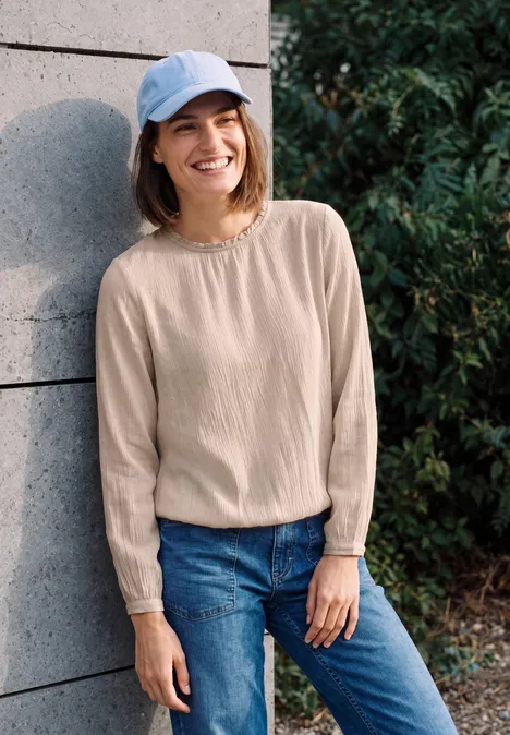Jeune femme en casquette bleue, pull beige et jean, appuyée contre un mur à l'extérieur, souriante.