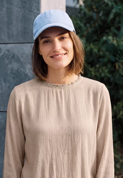 Une femme souriante porte un haut beige et une casquette bleu clair, devant un mur et des arbustes.