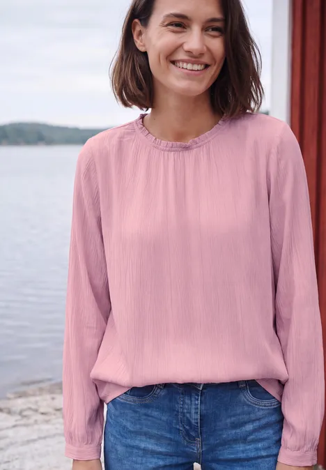 Langarm Bluse mit Rundhals und Smokdetail mellow rose Eine lächelnde Frau in einer rosa Bluse und Jeans am Ufer mit Wasser und Holzpfosten im Hintergrund.
