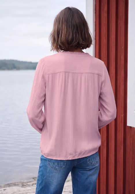 Langarm Bluse mit Rundhals und Smokdetail mellow rose Person steht mit dem Rücken zum Betrachter, trägt rosa Bluse, lehnt an roter Holzwand am Wasser.