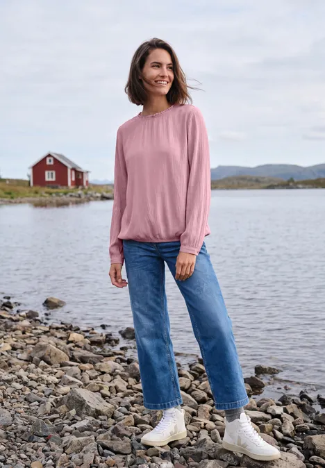 Langarm Bluse mit Rundhals und Smokdetail mellow rose Frau in Jeans und rosa Pullover steht am Ufer vor See und roter Hütte.