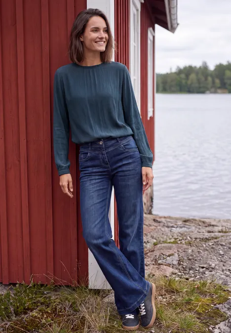 Blouse à manches longues et col rond avec détails smockés moonlight blue Une femme souriante en jean et pull bleu s’appuie détendue contre une maison rouge près d’un lac.