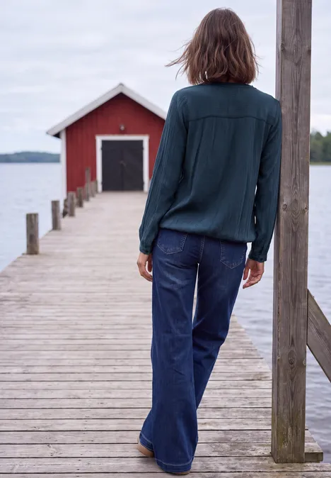 Blouse à manches longues et col rond avec détails smockés moonlight blue Une personne debout sur une jetée en bois regarde un abri à bateaux rouge au bord d’un lac.