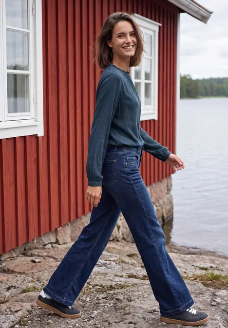 Blouse à manches longues et col rond avec détails smockés moonlight blue Une femme en jean et pull marche en souriant près d'une maison rouge au bord de l'eau.