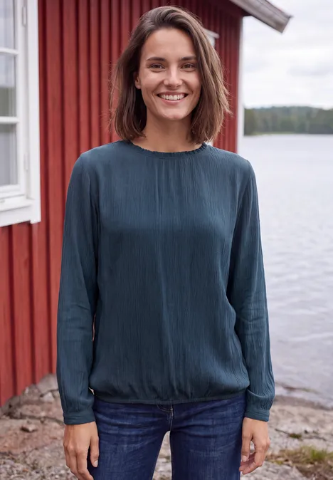 Blouse à manches longues et col rond avec détails smockés moonlight blue Femme souriante en chemisier bleu devant une maison rouge au bord d’un lac.