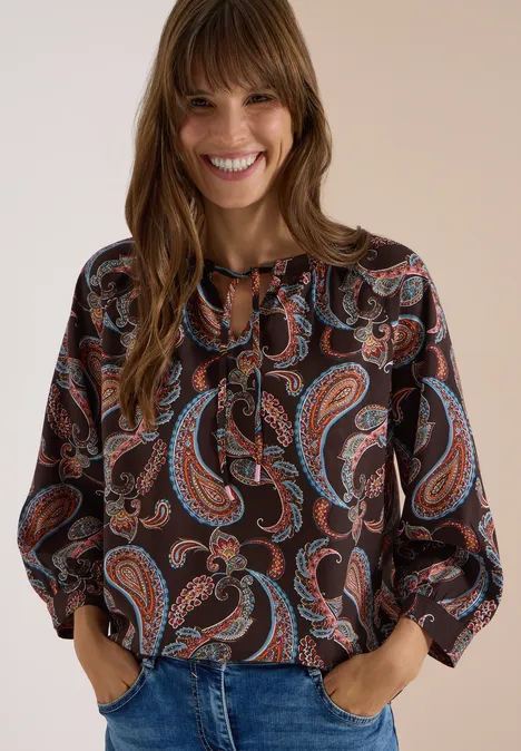 Frau mit lächelndem Gesicht, trägt eine bunte Paisley-Bluse und Jeans.