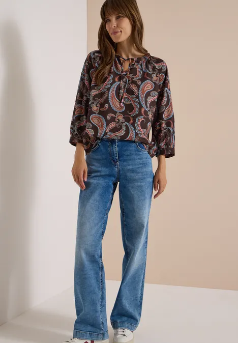 Frau in weicher Paisleybluse und weiten Jeans steht vor cremefarbenem Hintergrund.