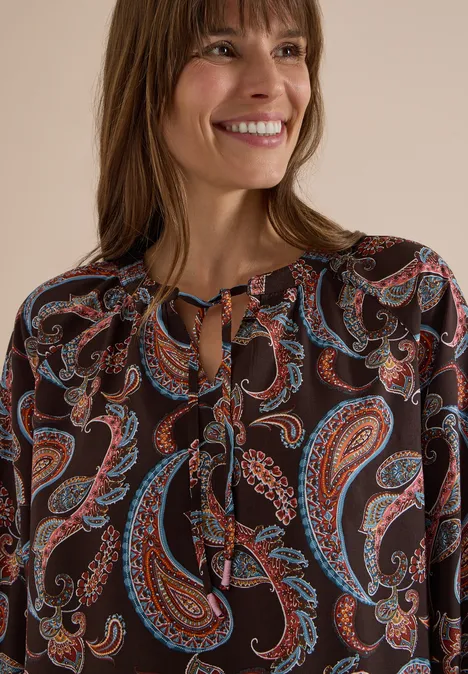 Eine lächelnde Frau trägt eine schwarze Bluse mit buntem Paisley-Muster vor hellem Hintergrund.