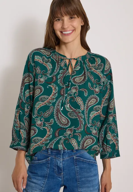 Frau mit grüner Paisley-Bluse und Jeans vor weißem Hintergrund.