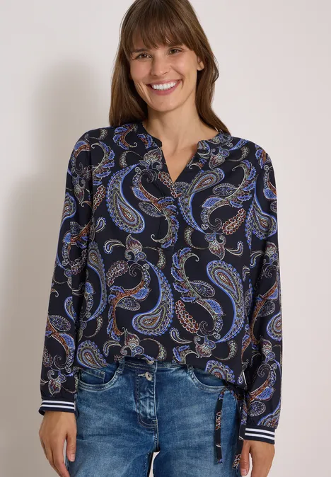 Eine lächelnde Frau in dunkler Paisley-Bluse und Jeans.