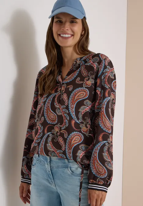 Vrouw met pet draagt spijkerbroek en lange mouwenblouse met kleurrijk paisleypatroon, lachend.