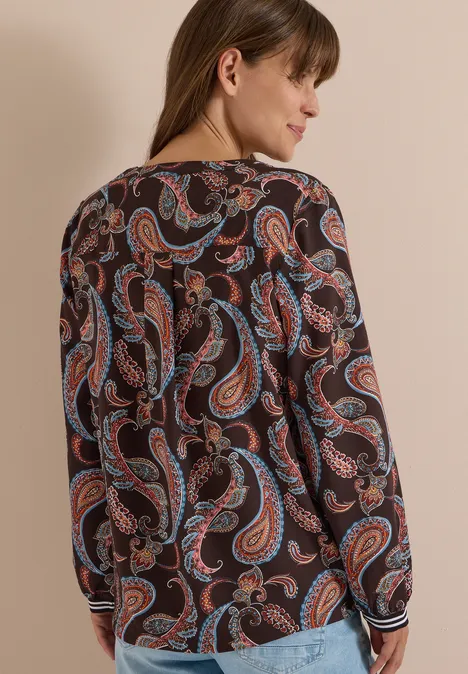 Vrouw van achteren in een bloes met lange mouwen en kleurrijke paisleyprint tegen neutrale achtergrond.