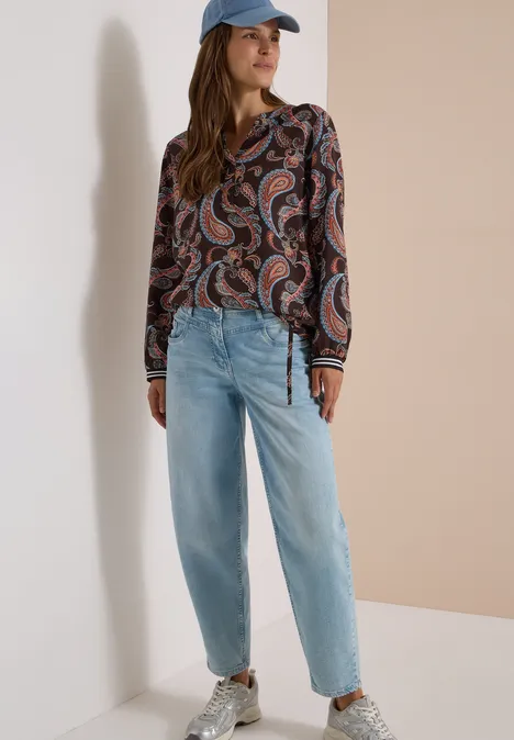 Vrouw in paisley-blouse, lichtblauwe jeans, witte sneakers en pet, leunend tegen een muur.