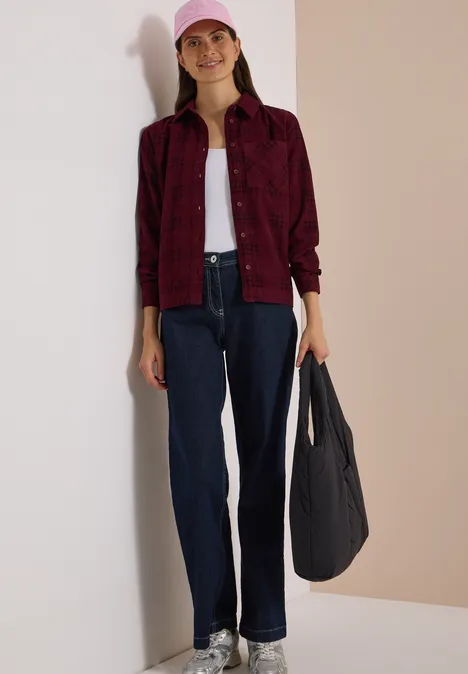 Frau in rotem Karohemd, weißem Top, dunklen Jeans, rosa Cap und schwarzer Tasche vor hellem Hintergrund.