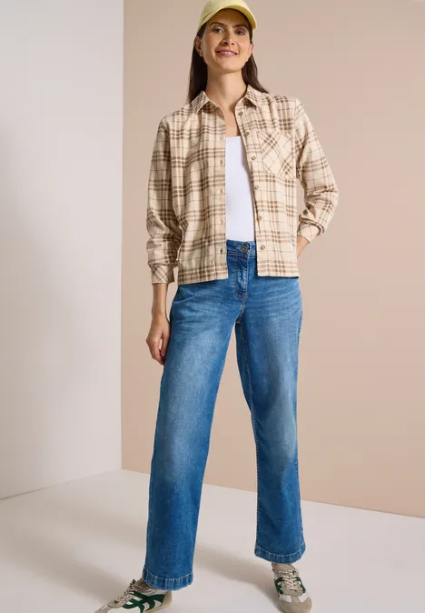 Frau in beiger karierten Hemdjacke, weißem Top, blauen Jeans und gelber Kappe vor neutralem Hintergrund.