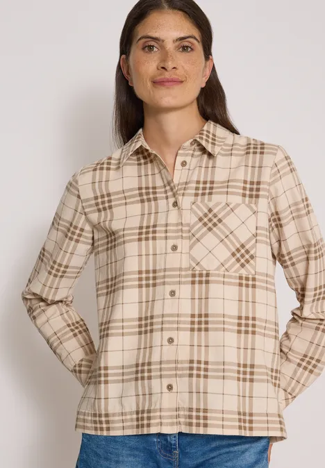 Frau trägt beige kariertes Hemd mit Brusttaschen und Jeans vor hellem Hintergrund.