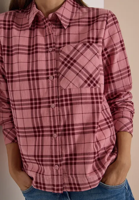Person trägt pink kariertes Hemd mit Brusttasche, Hände in Jeans-Taschen.