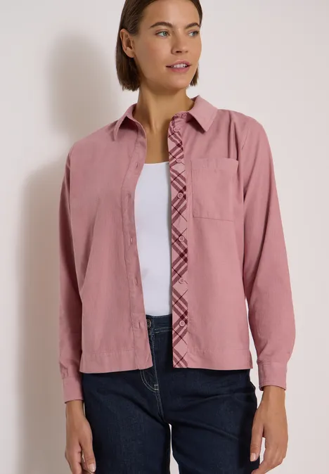 Frau trägt rosa Hemd über weißem Top und blaue Jeans, blickt seitlich