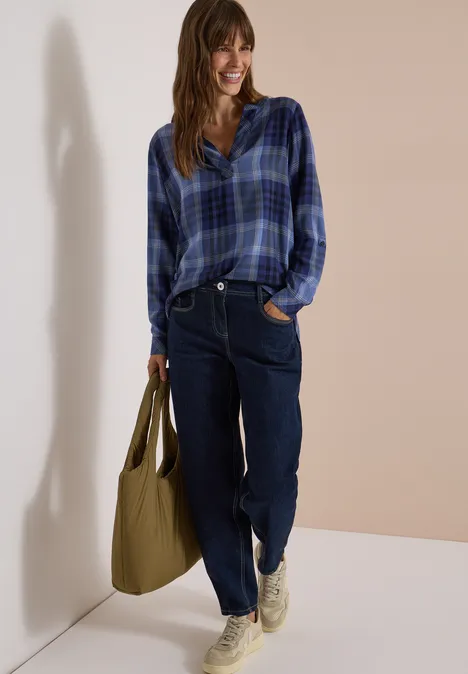 Modische Frau in blauem Karoshirt, dunkelblauer Jeans, weißen Sneakern und großer grüner Tasche.