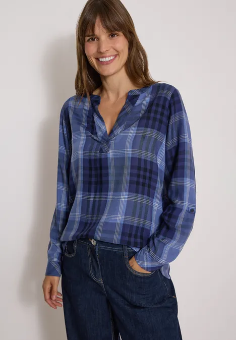 Frau in blau kariertem Hemd und blauen Jeans lacht vor hellem Hintergrund.